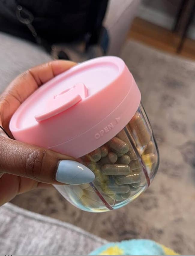 A hand holding a small clear container with a pink lid, filled with various capsules and pills