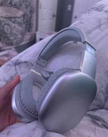 Silver over-ear headphones resting on a white blanket. One hand holds the headphones