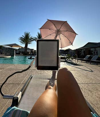 Person lounging by a pool reading an e-reader with a pink parasol providing shade; palm trees and lounge chairs in the background