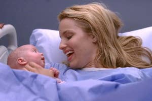 Dianna Agron, lying in a hospital bed, smiling and holding a newborn baby who is looking up at her as Quinn on Glee