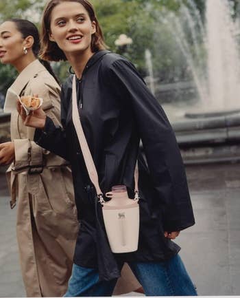 Two people walk in an urban setting; one holds a cone of food and wears a bag, casual coats, and jeans