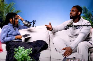 Two people are sitting on a couch engaged in a podcast conversation with microphones. One holds a tablet; the other gestures while speaking