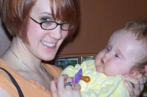 A woman with short hair and glasses smiles while holding a baby dressed in a onesie, who is looking at a pacifier in her hand