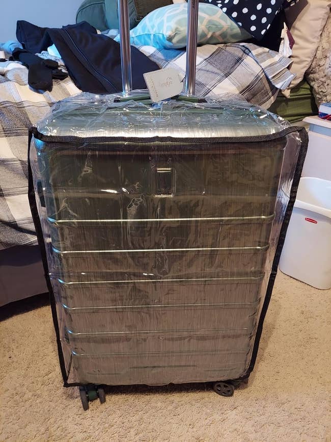 Large suitcase protected with a clear plastic cover, sitting in a bedroom with a bed and laundry basket visible in the background