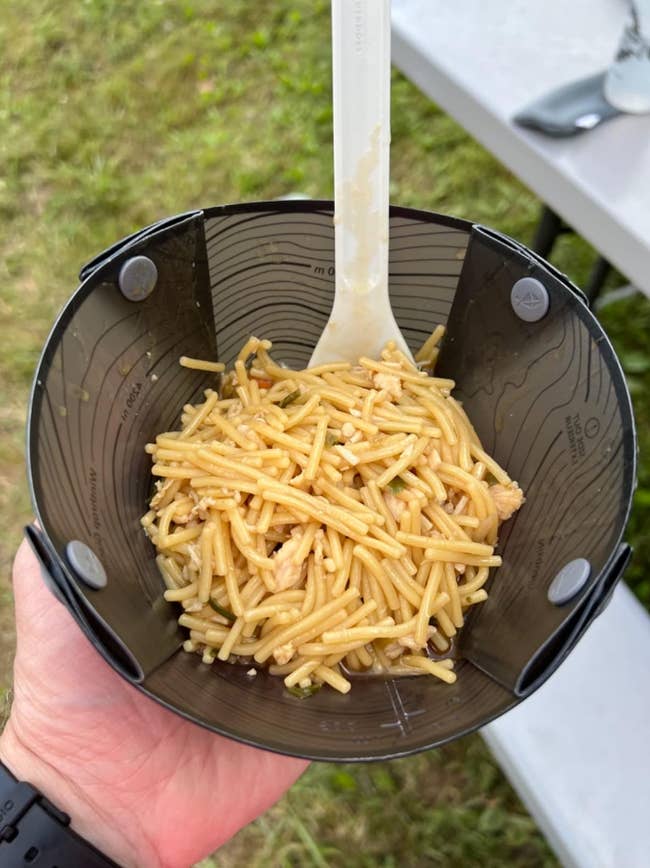 Spaghetti in a black camping bowl held by a hand, with a white spoon. Simple setup on a picnic table outdoors