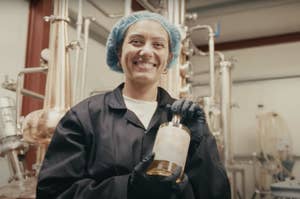 Person wearing a hairnet and gloves smiles while holding a bottle in an industrial setting