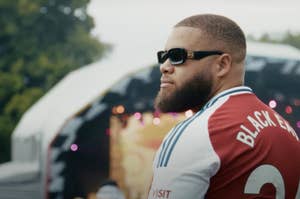 A person with sunglasses and a beard looks towards an outdoor stage, wearing a jersey with "Black 3" on the back