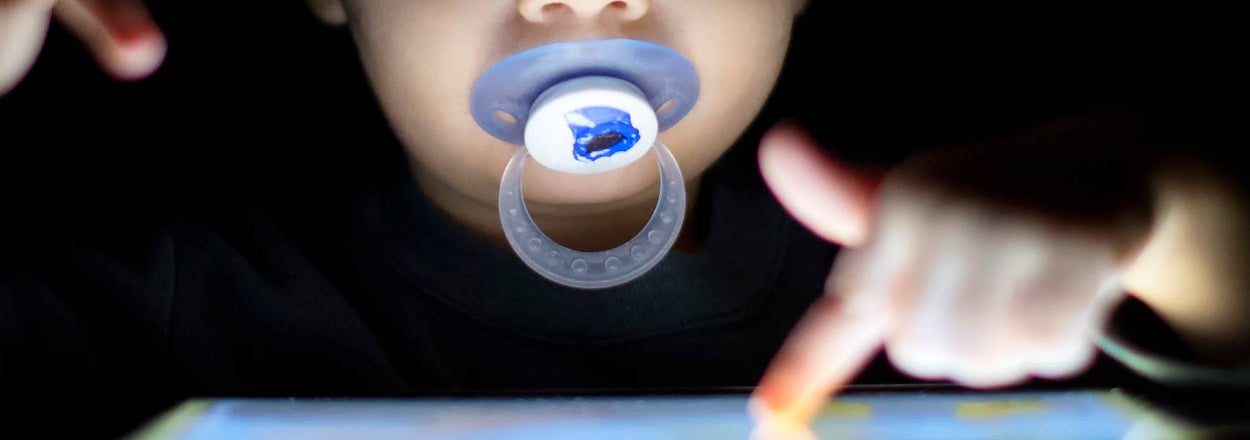 A child using a tablet in the dark with a pacifier in their mouth, engrossed in an activity on the screen