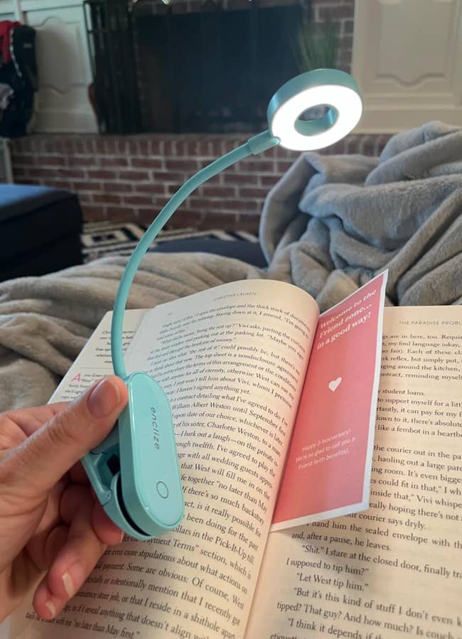 A hand holds a clip-on reading light illuminating an open book on a cozy blanket