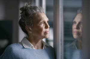 A woman with gray hair in a bun and wearing a white shirt under a light knit sweater looks thoughtfully out a window, her reflection visible in the glass