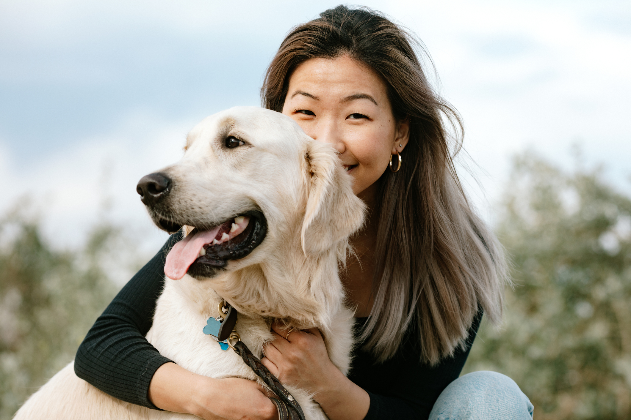 A woman smiles while holding a Golden Retriever. Both look happy outdoors