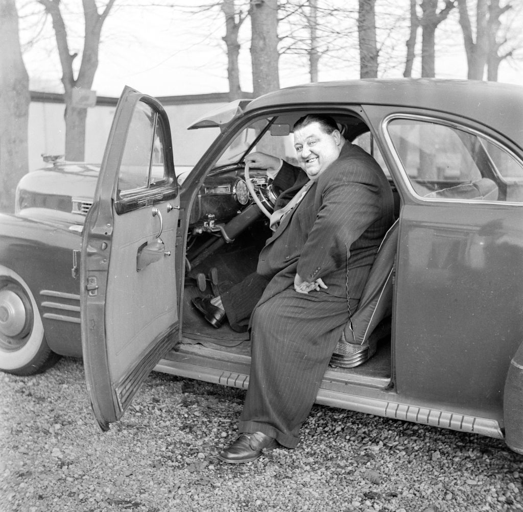 A man in a suit sits in the driver&#x27;s seat of an open car door while smiling at the camera