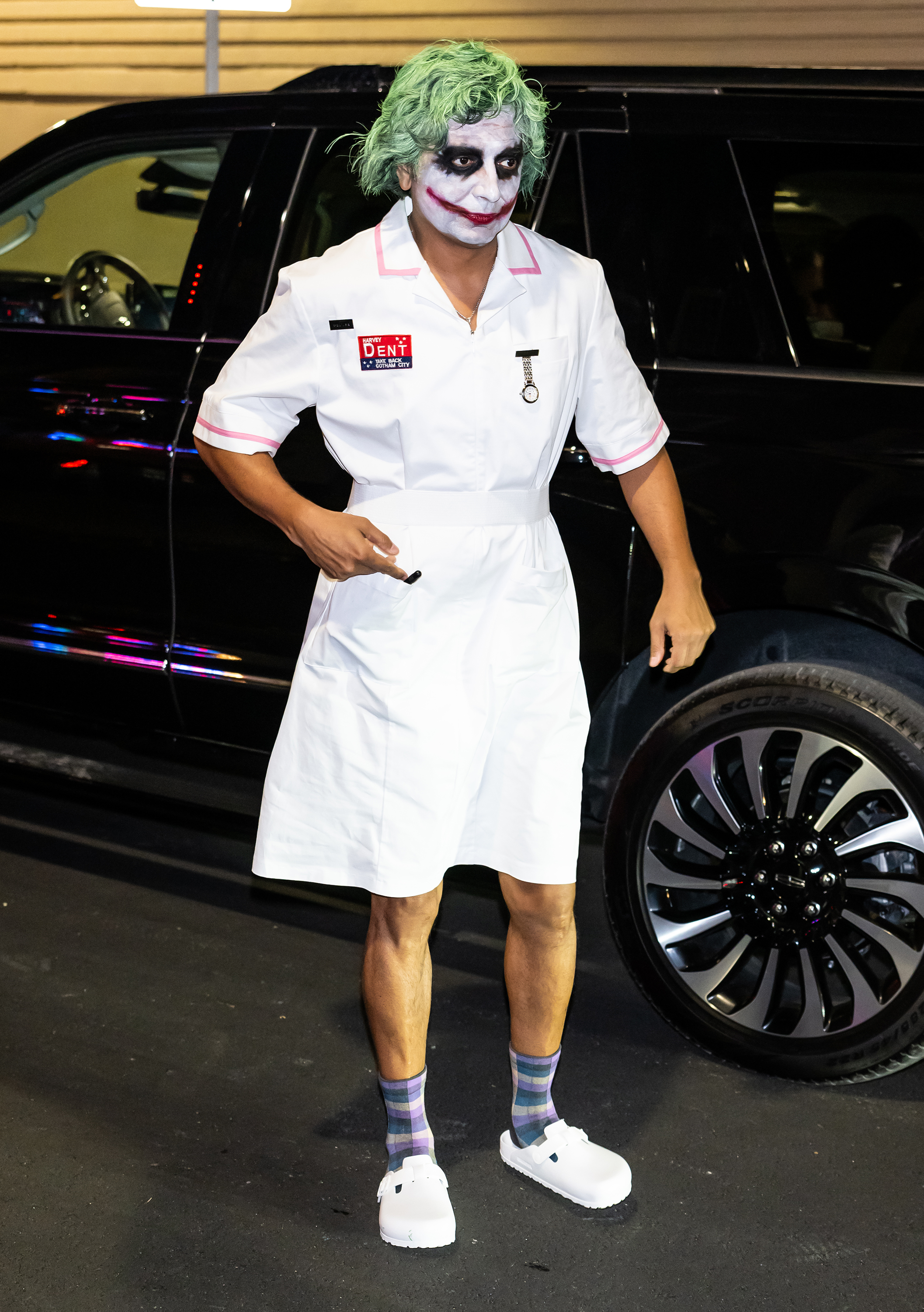 M. Night Shyamalan dressed as Joker wearing a nurse costume, Crocs, and striped socks in front of a vehicle