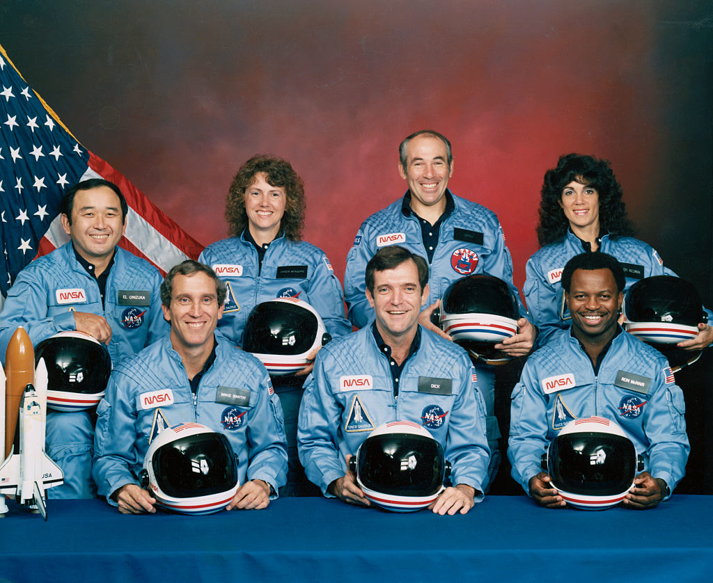 Seven NASA astronauts in blue flight suits pose with helmets, smiling. An American flag and a space shuttle model are in the background