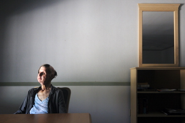 A person sits alone at a table in a dimly lit room with a mirror and a small bookshelf in the background