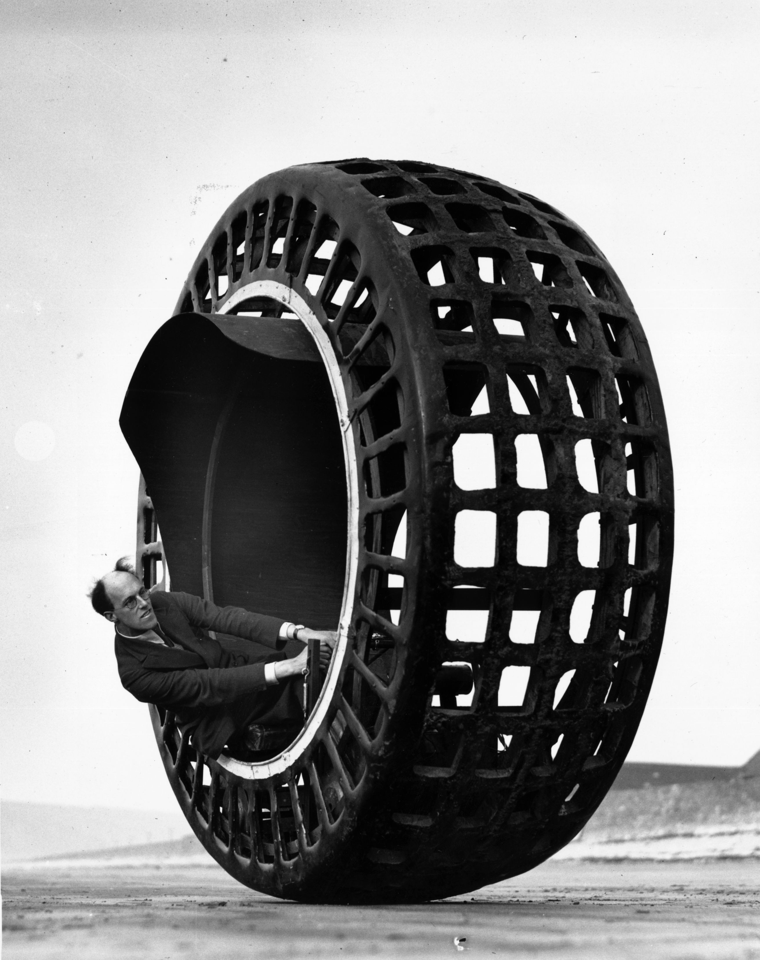 Man in a vintage mechanical device, resembling a large wheel with an open grid structure, seated inside and holding the handles