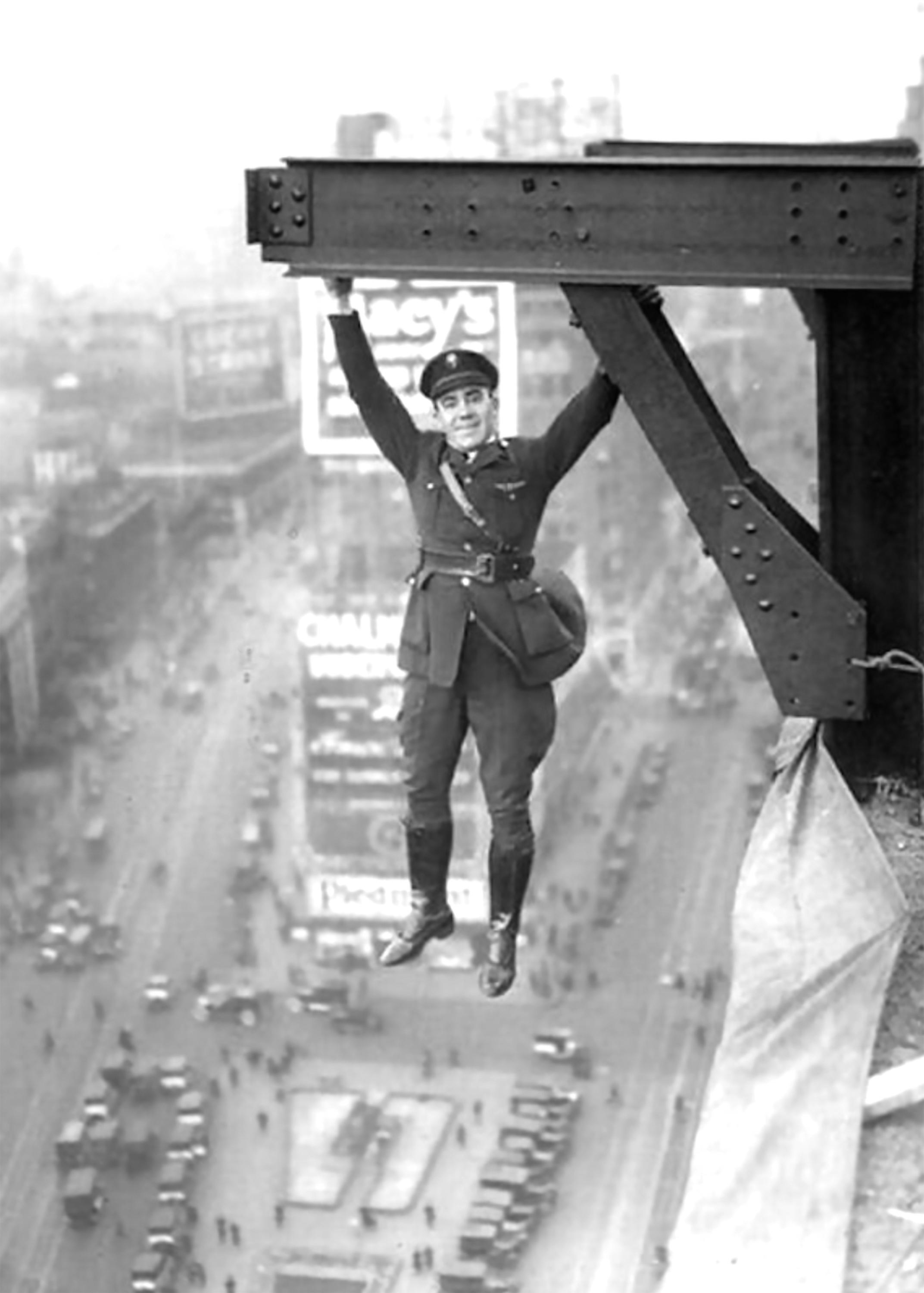 A man in a uniform hangs from a steel beam high above a busy city street in an old photograph