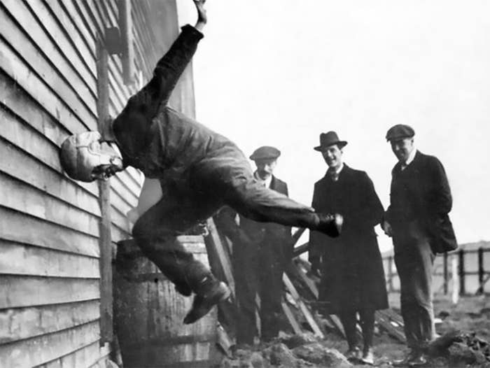 A black-and-white image showing a man diving headfirst into a wooden wall, observed by three men wearing suits and hats