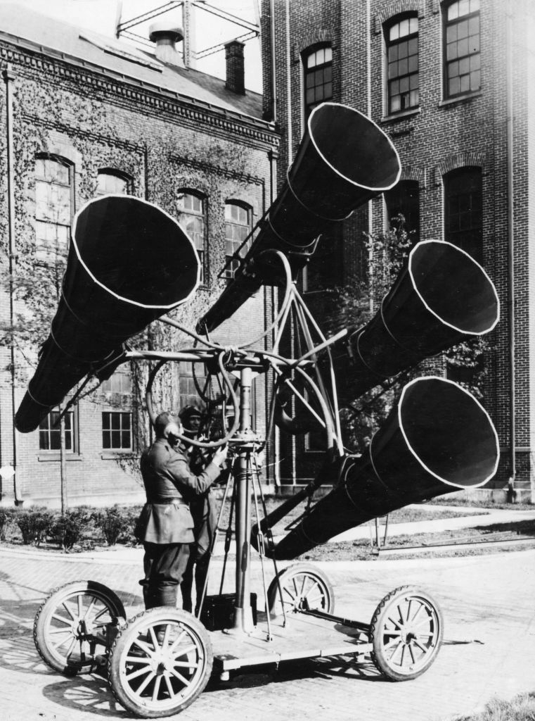 A person operates an early 20th-century acoustic locator, with large funnels on a wheeled base, in an industrial setting