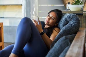 Woman in workout attire lounges on a cushioned chair, looking at her phone and wearing a fitness tracker