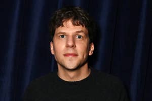 Jesse Eisenberg standing in front of a dark curtain, wearing a casual black sweater