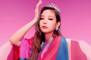 Jenny poses with a hand on their head, wearing a tiara and colorful outfit, sitting against a vibrant backdrop