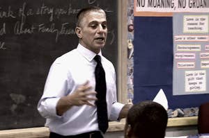 Tony Danza in a formal shirt and tie is speaking in front of a classroom. Behind him is a blackboard with notes and a poster with rules