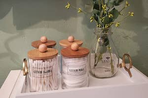 a reviewer photo of the labeled glass jars with wood lids filled with q-tips and cotton pads on a tray with a vase of eucalyptus