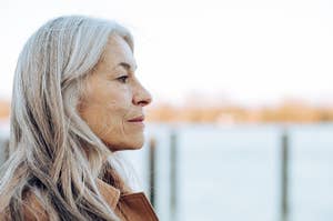 An older woman with long white hair looks thoughtfully into the distance by a waterfront