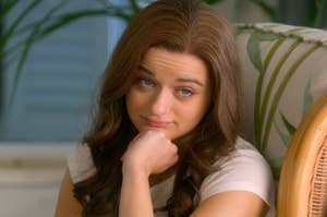 Joey King, wearing a short-sleeve top, rests her chin on her hand, looking contemplative in a casual indoor setting