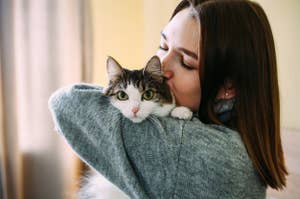 A woman kisses a cat while holding it close. The cat has white fur with darker markings and is looking directly at the camera