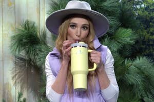 Chloe Fineman in a hat and sleeveless jacket sips from a large yellow mug through a straw, surrounded by pine trees