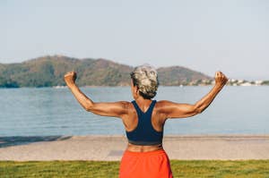 A person with short gray hair stands by a lake, flexing their muscles while facing the water and distant hills