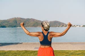 A person with short gray hair stands by a lake, flexing their muscles while facing the water and distant hills