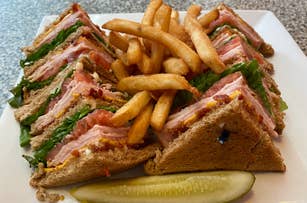 Club sandwich with turkey, bacon, lettuce, and tomato, served on wheat bread with a side of french fries and a pickle slice