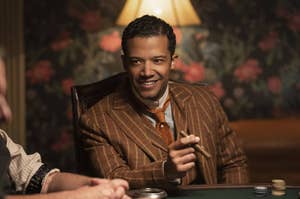 Jacob Anderson in a patterned suit sits at a table with poker chips, smiling and holding a cigar